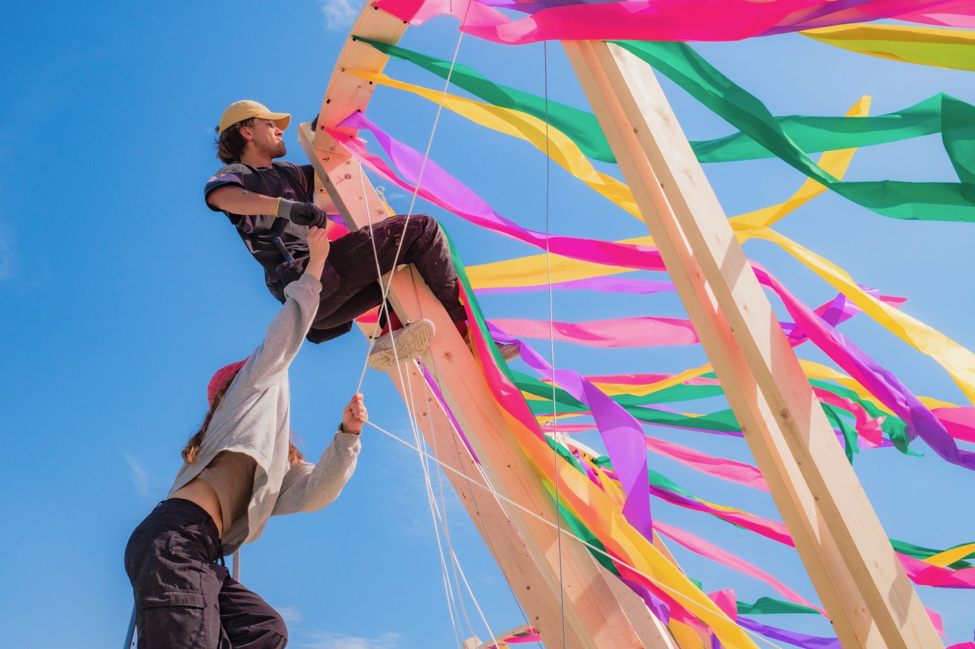 Two people assembling a timber arch hung with pink, yellow, green and purple ribbons against a blue sky